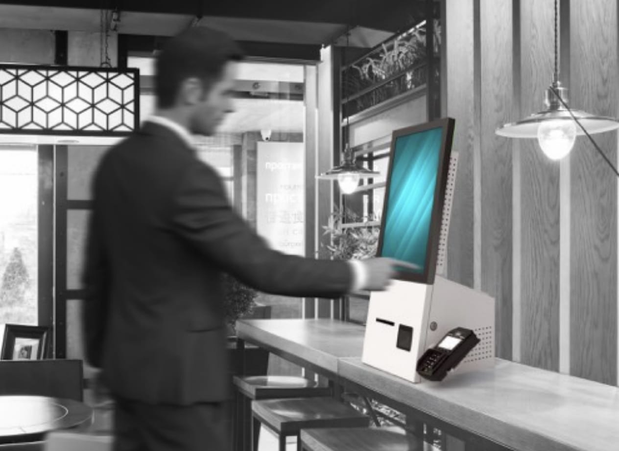 Un homme en costume interagit avec un kiosque libre-service à écran tactile K2 CORP sur un comptoir dans un cadre intérieur moderne.