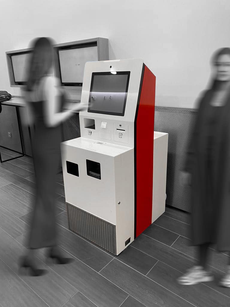 Deux femmes se tiennent près d'un kiosque libre-service à écran tactile avec une bande rouge dans un cadre intérieur moderne ; l'arrière-plan et les personnages sont flous.