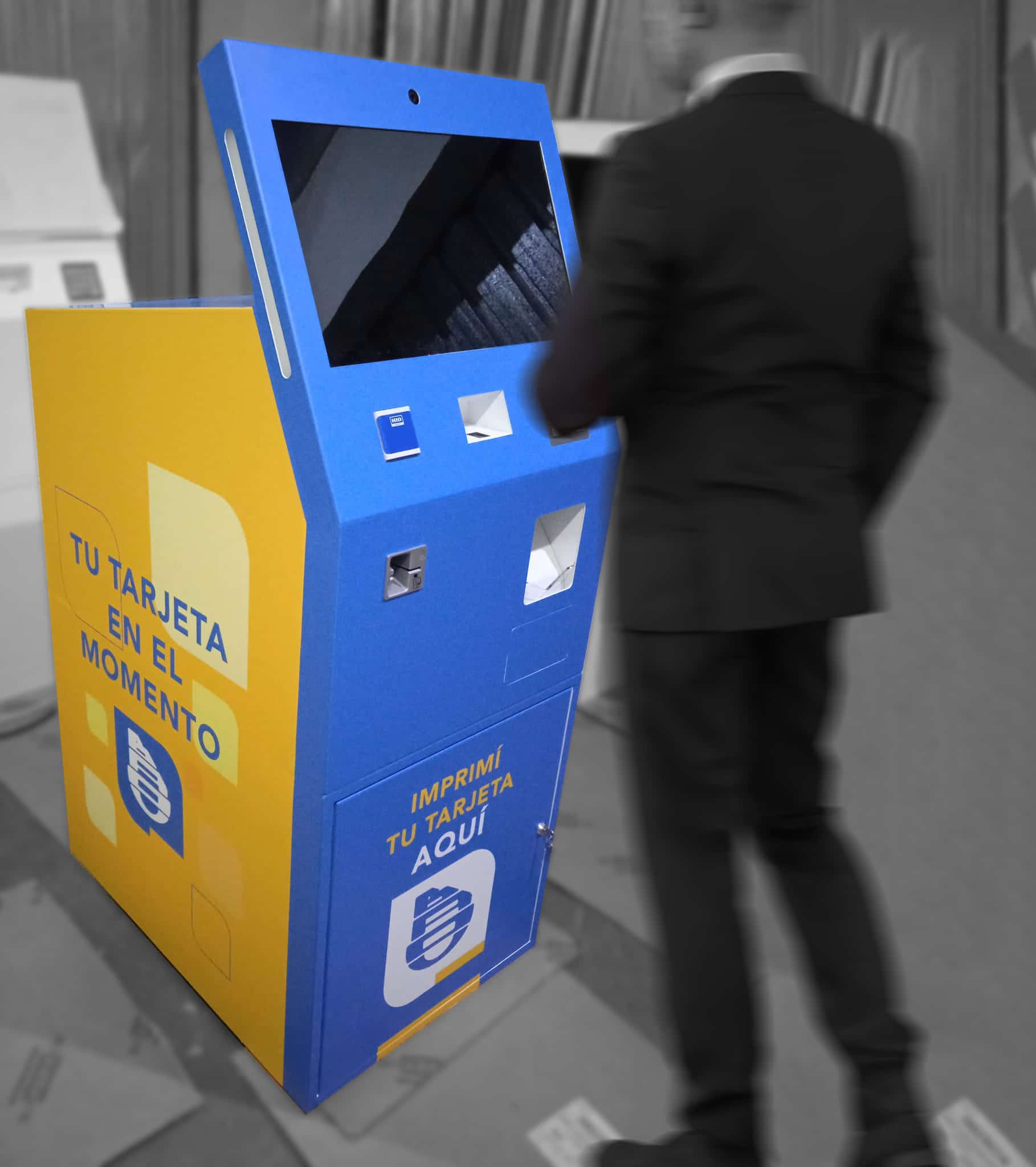 Un homme en costume se tient devant un kiosque libre-service bleu et jaune avec un texte espagnol pour l'impression instantanée de cartes.